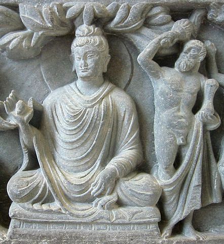 A high relief engraving made of gray stone, showing the Buddha in Lotus posture in a robe with a halo and bun, upholding something in His right hand. To His left is what looks like Heracles, nude except for a cloth that drapes below His knees, holding what looks like a club over one shoulder and some other object in His left hand.