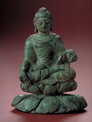A figurine of the Buddha with a dark green patina. He is seated cross-legged on a double lotus, wearing a long flowing robe and holding up His left hand.