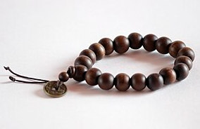 A closeup of a wrist mala. It is made of 18 perfectly spherical beads of dark brown wood. The final guru bead has a small bead and cash coin strung on a tassle of two black strings.