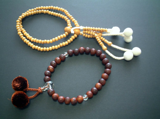 Two Japanese Buddhist juzu. The top one is a double juzu made with small, light straw color beads and white pompoms. The bottom one is a single bracelet of larger, dark brown beads only 28 in number (with two, clear glass counter beads) and a clear glass Meru bead that has two chocolate colored pompoms at the end.