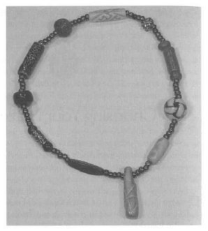 A grainy black and white photo of a circular necklace of beads. Ten large beads alternate between different sized circular and cylindrical shapes with five tiny seed beads separating each one. There is an eleventh cylindrical bead at the end, hanging like a pendant.