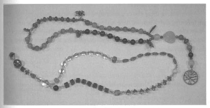 A grainy black and white photo of two long, beaded necklaces that are shaped like Christian rosaries. In place of the crucifix is a Tree of Life. The counter beads are replaced with metal charms of things like pinecones and feathers. Each decade has a different color or shape of bead.