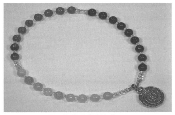A grainy black and white photo of a small bracelet. The beads are all perfectly round, glass beads divided into three groups of nine. Each group has a different color. Small clear seed beads separate the main beads. The bracelet ends with a medal that has a spiral design on it.