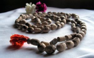 Closeup of a tulsi bead mala in soft white lighting. The beads are rough hewn, rounded cylinders in a soft, pale wood, with a matte texture. The final meru bead has an orange-red tassle at the end