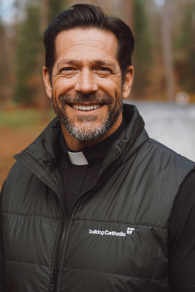 A handsome white man with combed back brown hair, perhaps in his 40's, whose face could be compared to Jon Hamm's. His smile shows his pearly white teeth. He wears a clerical collar under his sporty Patagonia style vest that has "bulldog Catholic" stitched on it with a cross on a shield.