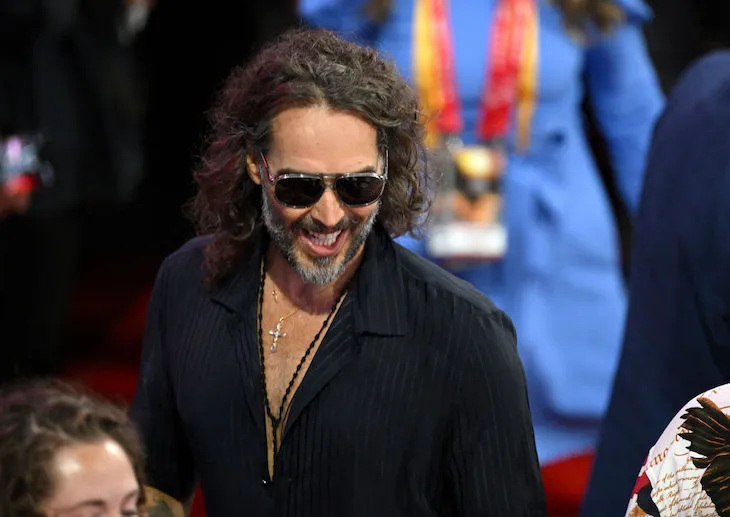 Russell Brand with a giant, toothy grin, wearing large sunglasses and a diamond cross necklace