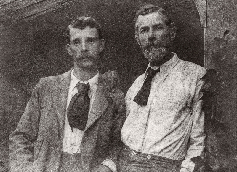 A black and white photo of two men leaning against each other and a doorway. On the left is a younger man with a large mustache and oversized necktie that is cut off at half length. On the right is an older man with a beard, resting his hand on the other's shoulder. He also has a necktie that appears cut short.