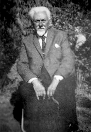A black and white photo of a man sitting outdoors in a suit and tie. His white hair is combed back and he sports a white mustache and goatee.