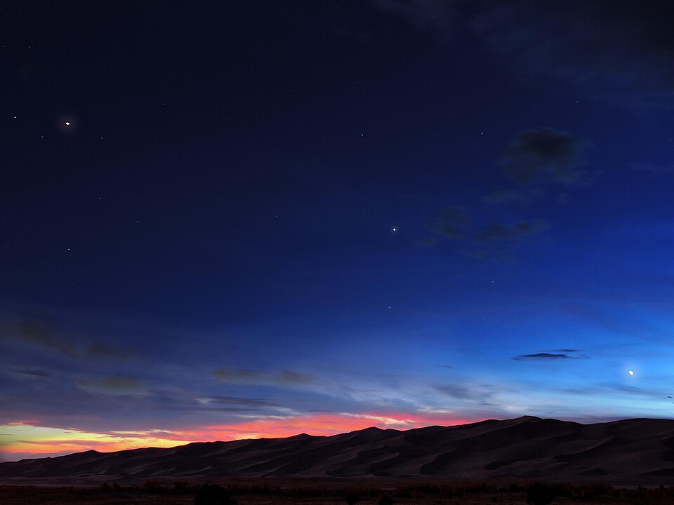 A deep blue night sky with a fiery hint of sunrise on the horizon, behind dark sand dunes. In a diagonal line across the sky (from top-left to bottom-right) are three large, white stars: Mars, Saturn, and Venus.