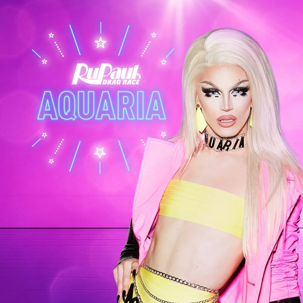 A promotional photo the drag queen Aquaria on Ru Paul's Drag Race. She is wearing a blonde wig, tiny yellow crop top, and hot pink coat, with a necklace that spells "Aquaria".